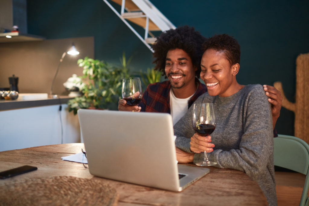 A millennial couple sitting in their kitchen and using their laptop to browse a winery website while drinking red wine.